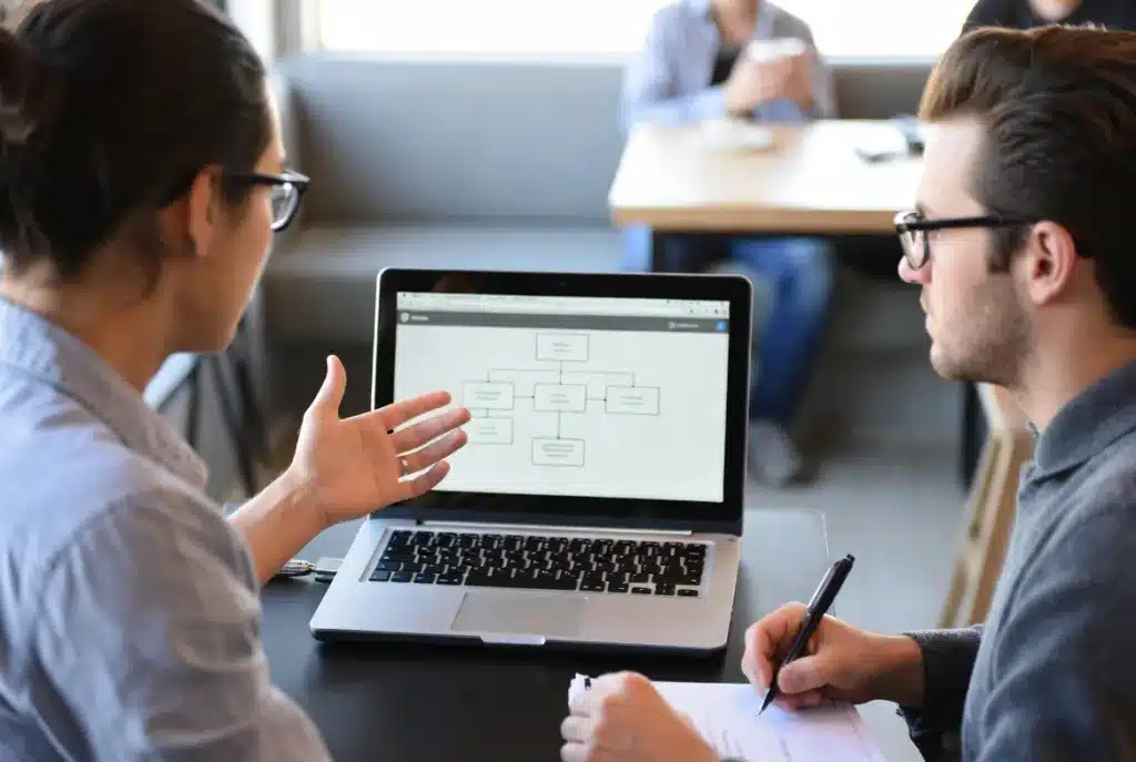 Two people in a discovery or strategy meeting. One person is presenting, the other is listening and taking notes. A laptop is open between them showing a website wireframe or sitemap. The setting feels consultative and professional, not salesy. A coffee shop or modern office.