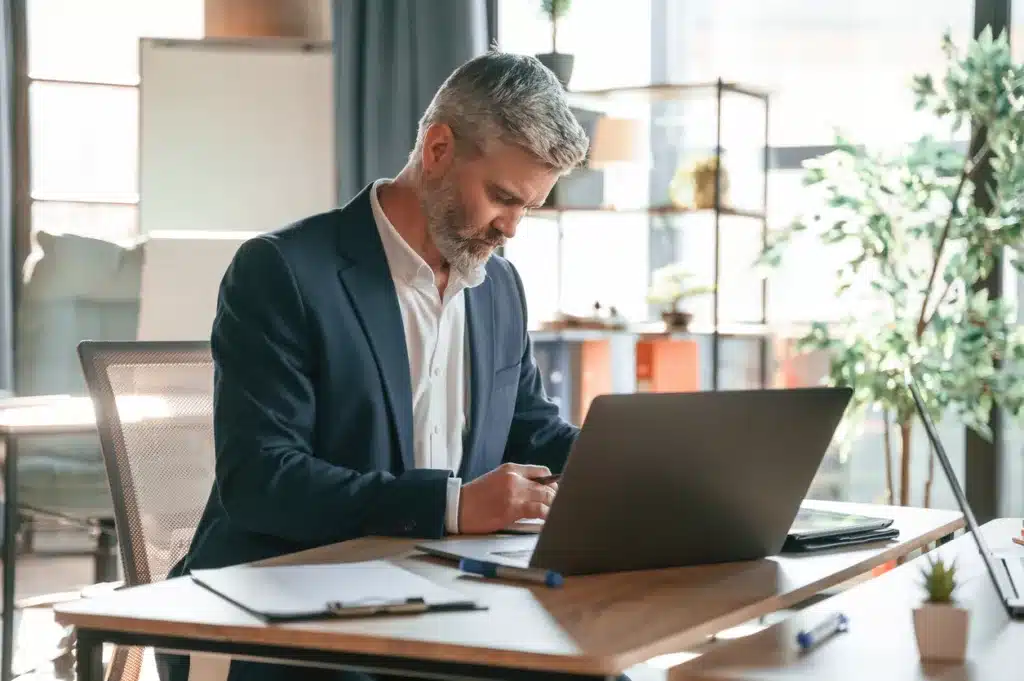 A businessman wearing a suit sits in a modern office, taking notes on his laptop about how to write a blog post.